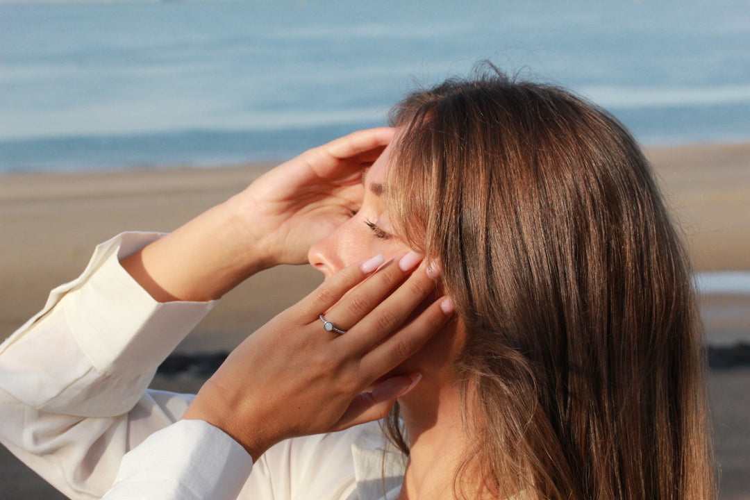 Een vrouw met lang bruin haar staart naar de oceaan en raakt haar gezicht aan met beide handen. Ze draagt een wit shirt en een sterling AsRing Zilver van See You terwijl ze op het strand staat, omringd door de serene zee en het zand.