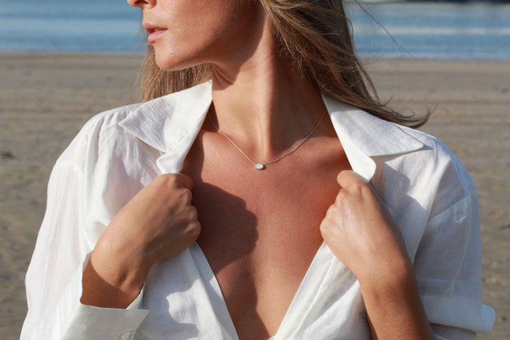 Op een rustig strand houdt een persoon de kraag vast van een open wit shirt en draagt hij Luna | AsKetting Zilver van See You, een elegante ketting met een klein rond hangertje, tegen de achtergrond van de oceaan en het zand.