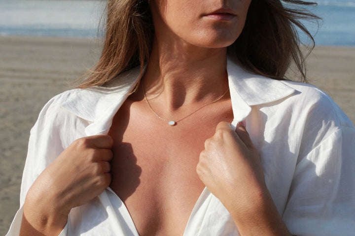 Op het strand draagt een persoon in een wit open shirt de delicate Luna | AsKetting Zilver van See You. Ze houden hun shirt met beide handen vast en kijken weg van de camera. Het zand en de oceaan vormen een perfecte achtergrond voor hun minimalistische klassieke ovale vorm ensemble.