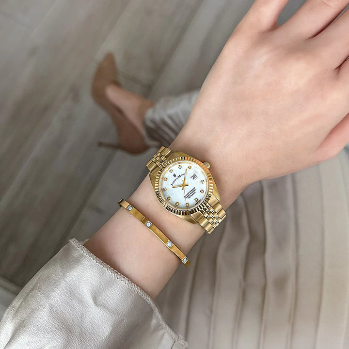Een persoon die de Jacques Du Manoir Inspiration 31mm met witte wijzerplaat en gouden armband draagt, paart deze met lichtgekleurde kleding en beige hoge hakken terwijl ze op een lichte houten vloer staat.