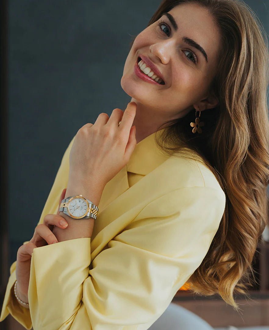Een vrouw met lang bruin haar, in een gele blouse en bloemetjesoorbellen, lacht naar de camera. Ze laat haar kin op haar hand rusten en toont de stijlvolle Jacques Du Manoir Inspiratie klassieke dameshorloge.