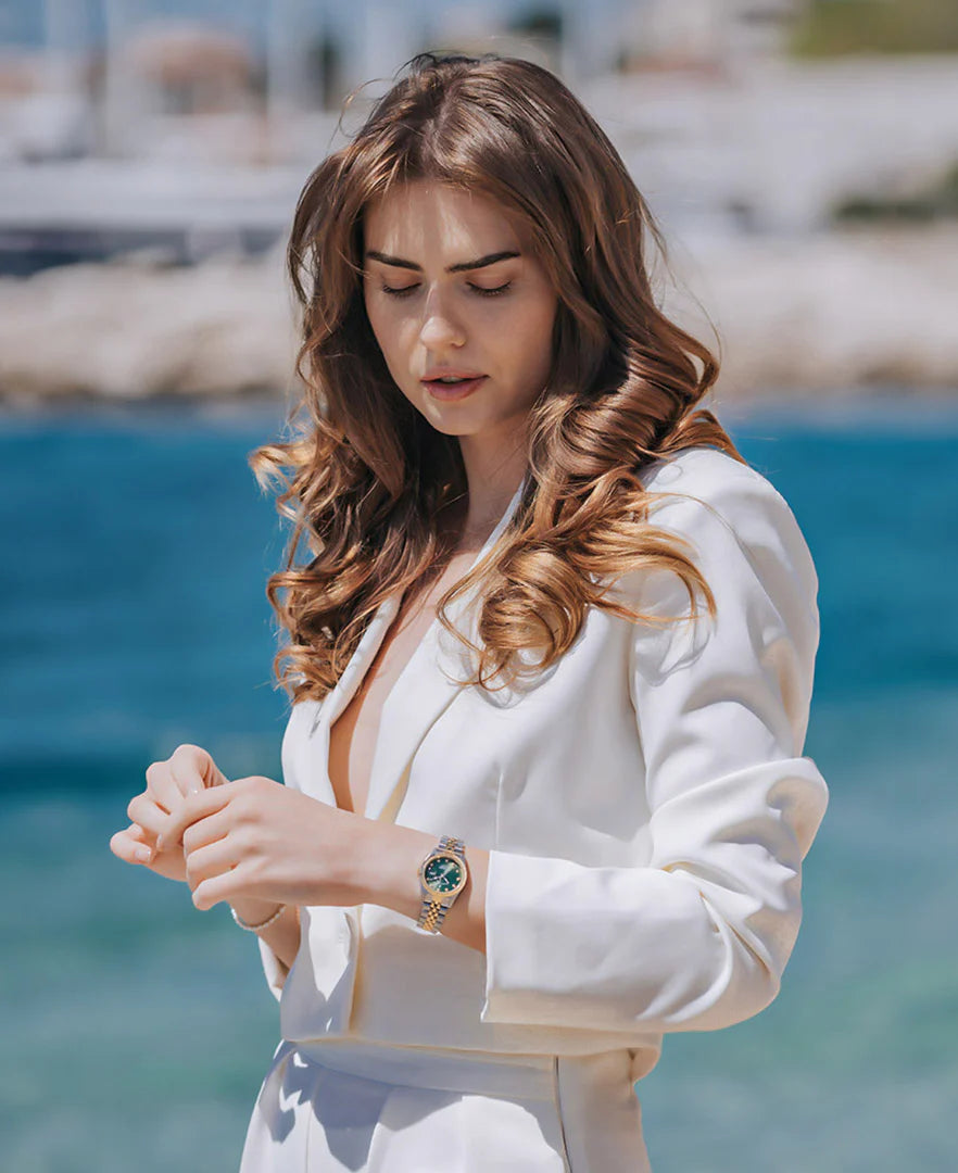 Een vrouw met lang, golvend bruin haar in een wit pak staat bij het blauwe water, nadenkend kijkend op haar Jacques Du Manoir Inspiration 31mm horloge. De zachte onscherpe achtergrond suggereert een zonnige dag aan zee.