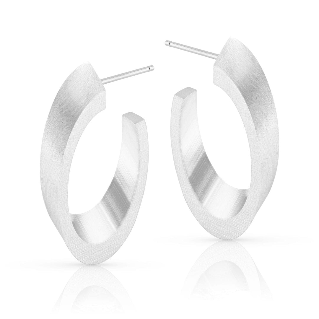 De Ovale Creolen O21 van CLIC Creations zijn zilveren creolen met een matte afwerking en open achterkant, met gladde, licht afgeplatte oppervlakken. Gemaakt van chirurgisch edelstaal, ze staan rechtop en worden weerspiegeld op een wit oppervlak.