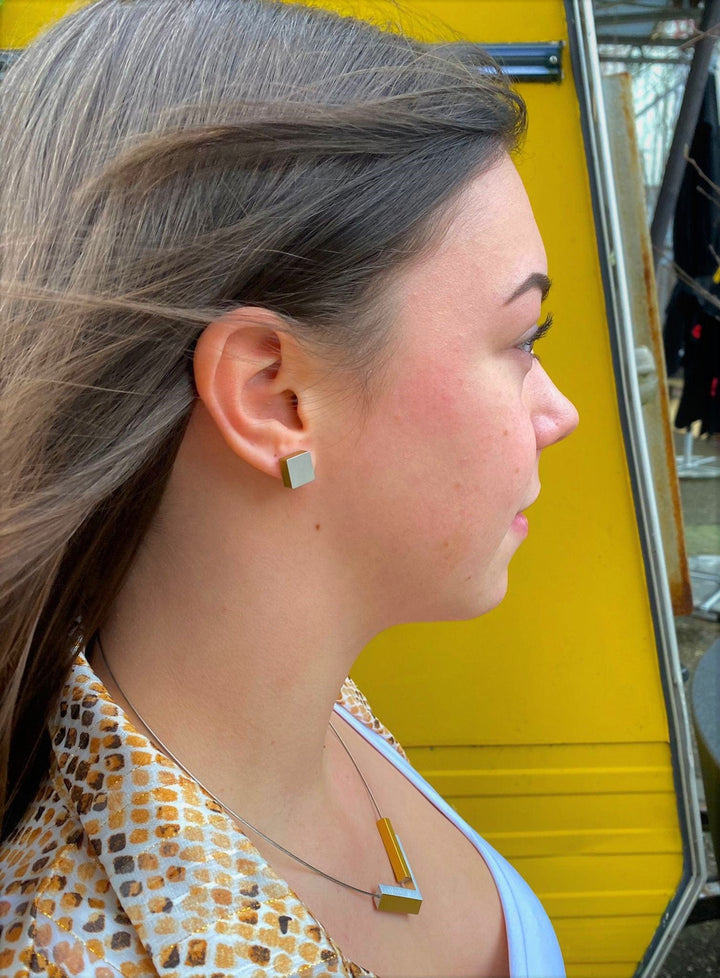 Een vrouw met lang, donker haar draagt CLIC Creations Oorsteker Vierkant Goud|Geel O15G oorbellen en een bijpassende hypoallergene aluminium ketting. Ze kijkt opzij, glimlacht lichtjes en toont haar uniek ontworpen accessoires tegen de felgele muur op de achtergrond.