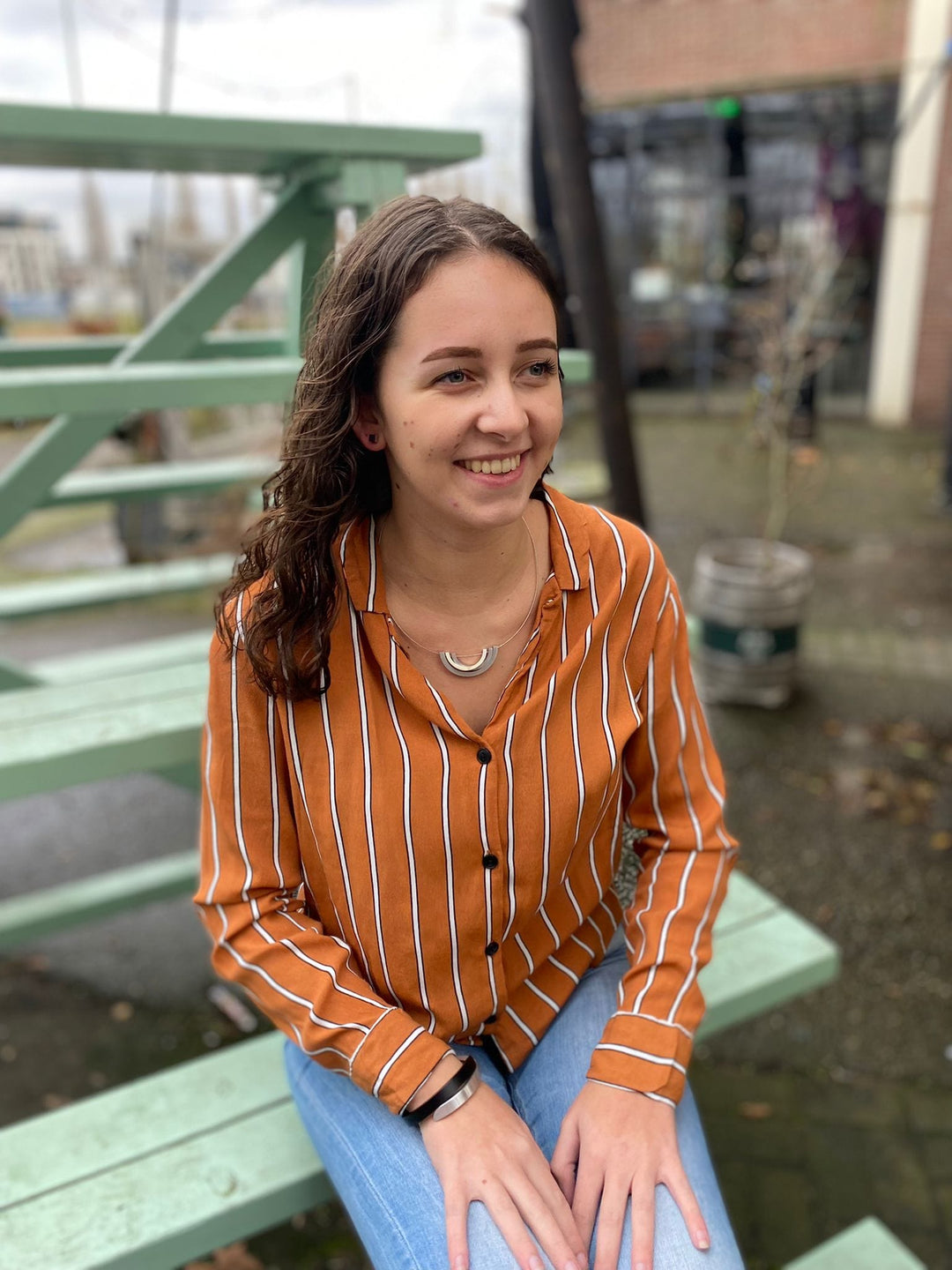 Een vrouw met lang bruin haar lacht op lichtgroene trappen, gekleed in een oranje en wit gestreept shirt en blauwe jeans. De scène roept het handgemaakt in Nederland gevoel op van CLIC Creations Armband Cs gesloten Oranje A1O terwijl ze zit te midden van een wazige buitenachtergrond met een gebouw en planten.