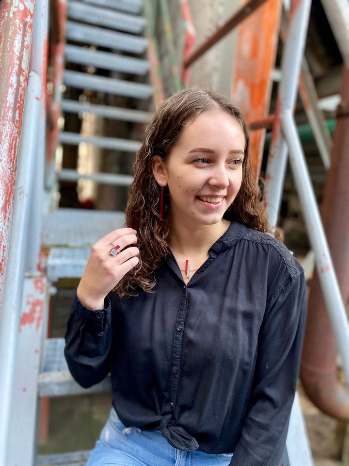 Een jonge vrouw met krullend haar lacht op een metalen trap, gekleed in een zwarte blouse, jeans, lange oorbellen en de Ring vierkant en kubusje Rood fits most R7R van CLIC Creations, die het Nederlandse vakmanschap benadrukt. De industriële setting omvat roestige rode en blauwe trappen op de achtergrond.