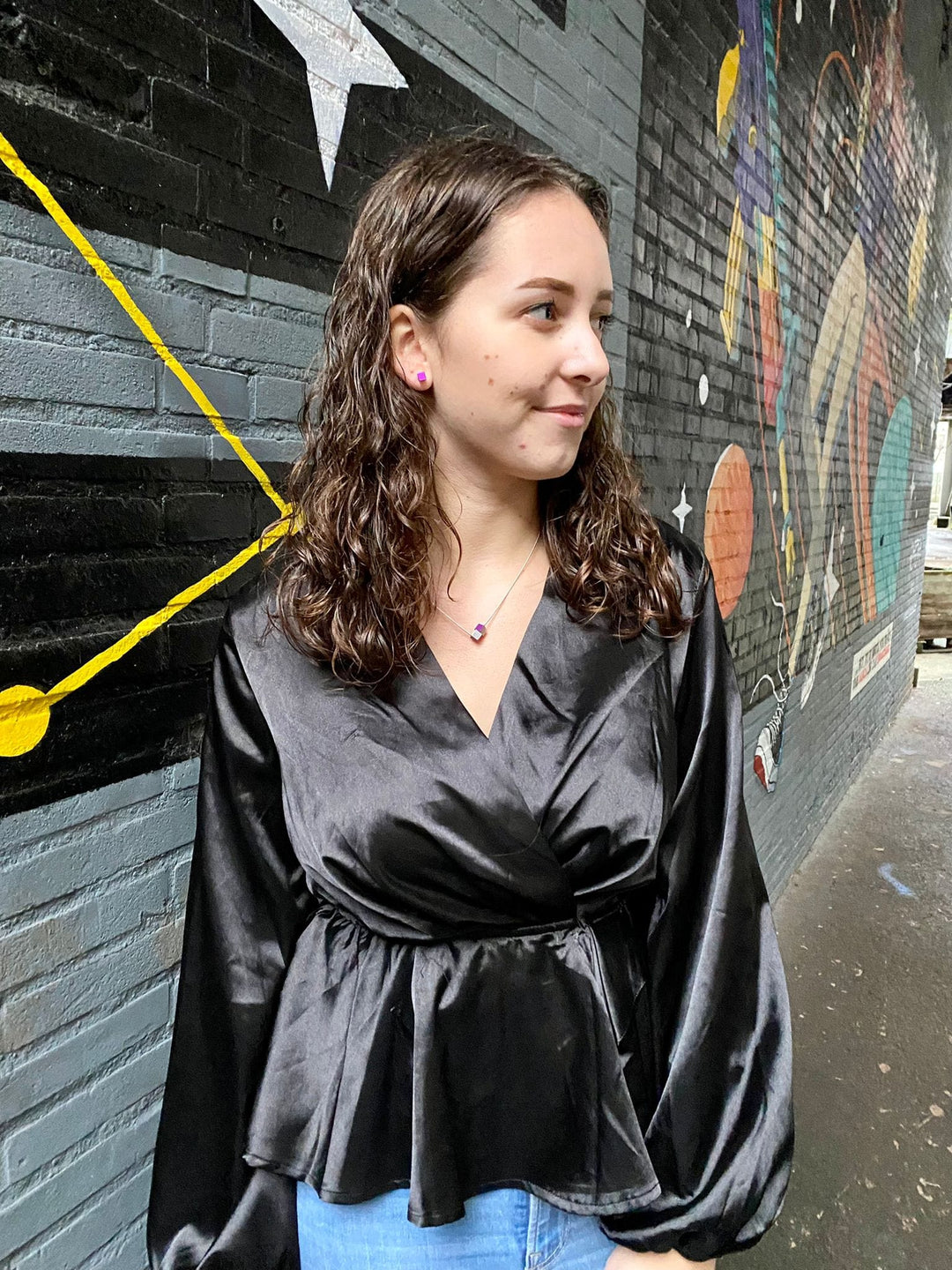 Een vrouw met krullend bruin haar, gekleed in een glanzend zwart topje, staat lachend naast een kleurrijke muurschildering van geometrische en abstracte ontwerpen die doen denken aan de handgemaakte Collier Twee kubusjes Oranje C205O van CLIC Creations.