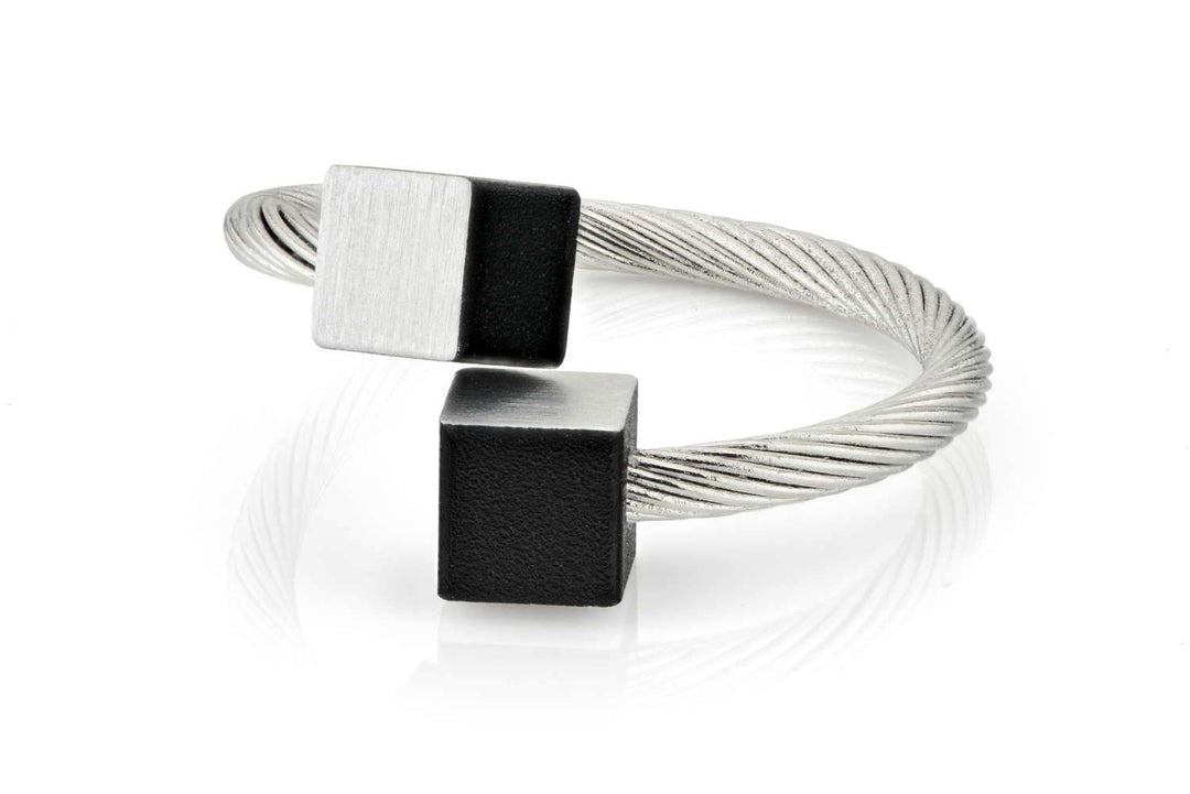 De Ring Kubusjes Zwart R4Z van CLIC Creations is een moderne, verstelbare ring van gedraaid chirurgisch staal met open uiteinden en vierkante accenten: één matzwart, de ander geborsteld zilver.