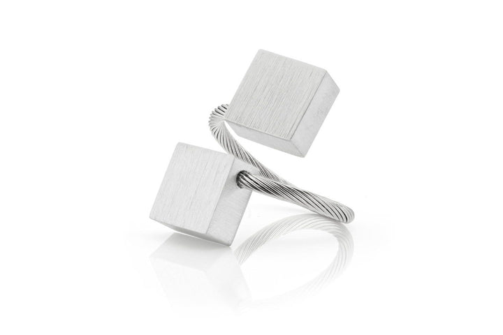 De Ring Vierkanten R6 van CLIC Creations is een minimalistisch metalen sculptuur, handgemaakt in Nederland. Het bestaat uit twee kubusvormige elementen die met elkaar verbonden zijn door gedraaide draad en heeft een zilveren afwerking, elegant gebalanceerd op een reflecterend oppervlak, wat lijkt op een perfect vervaardigde ring.