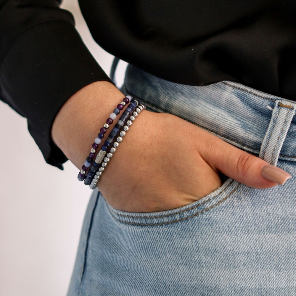 Een persoon in een zwart shirt met lange mouwen en een lichtblauwe spijkerbroek, met één hand in zijn zak, toont drie kralenarmbanden, waaronder de Rebel & Rose Silver Shine - 4mm Kralen Armband Dames in blauw, zilver en paars.