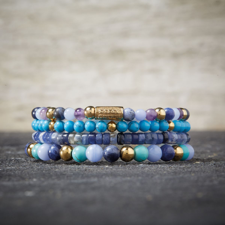 Four Rebel & Rose Zijn viooltjes blauw? 18 kt geelgouden geïoniseerde 4 mm kralenarmbanden voor vrouwen tonen blauwe, paarse, turquoise en gouden kralen die op elkaar gestapeld zijn op een donker gestructureerd oppervlak met een neutrale achtergrond.