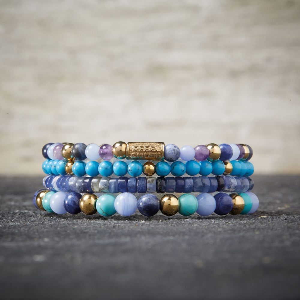 Een stapel kralenarmbanden van Rebel & Rose, met de Winter Blues - 6mm - Kralen Armband Dames, wordt weergegeven op een grijs gestructureerd oppervlak tegen een vage beige achtergrond in blauwe, turquoise, paarse en gouden tinten.