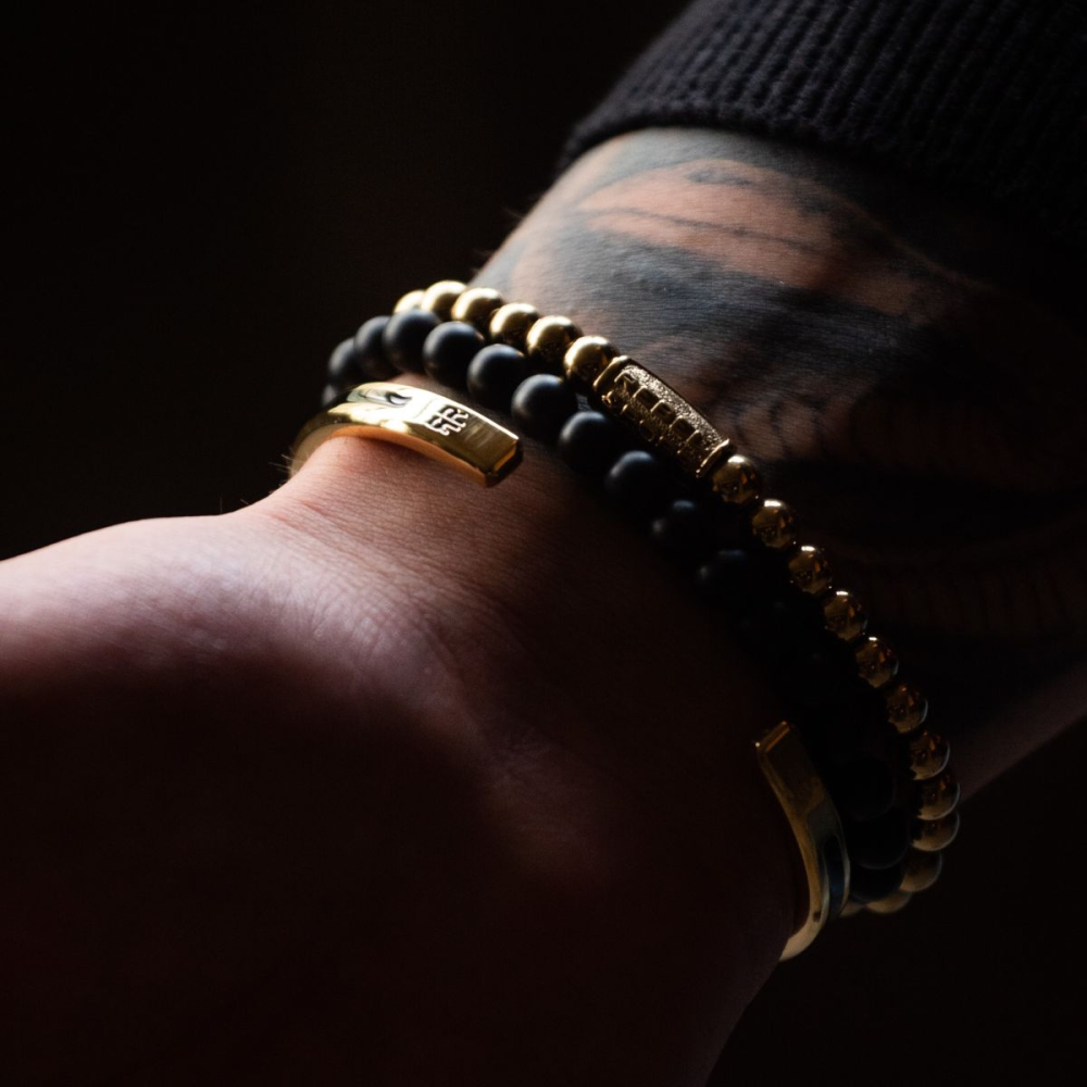 Een close-up van een getatoeëerde pols toont de Rebel & Rose Yellow Gold Only - 6mm Kralen Dames Armband die gloeit bij weinig licht, gestapeld met twee gouden armbanden en een zwarte kralenarmband die de 18kt ion-goud vergulde glans accentueren.