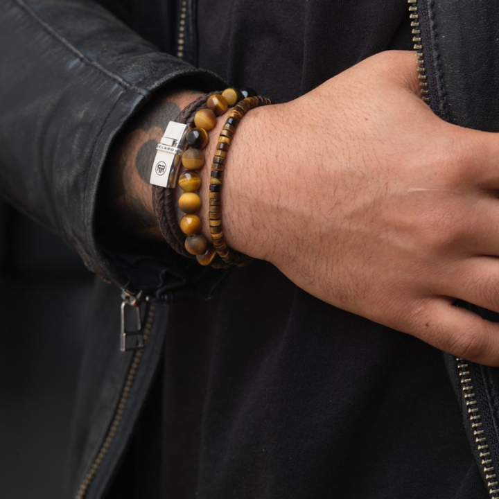 Een persoon met een zwart leren jack en shirt toont zijn pols versierd met meerdere aarde-tone heren armbanden, waaronder de Rebel & Rose Tiger Lily - 8mm Kralen Armband Heren met een klein metalen label.