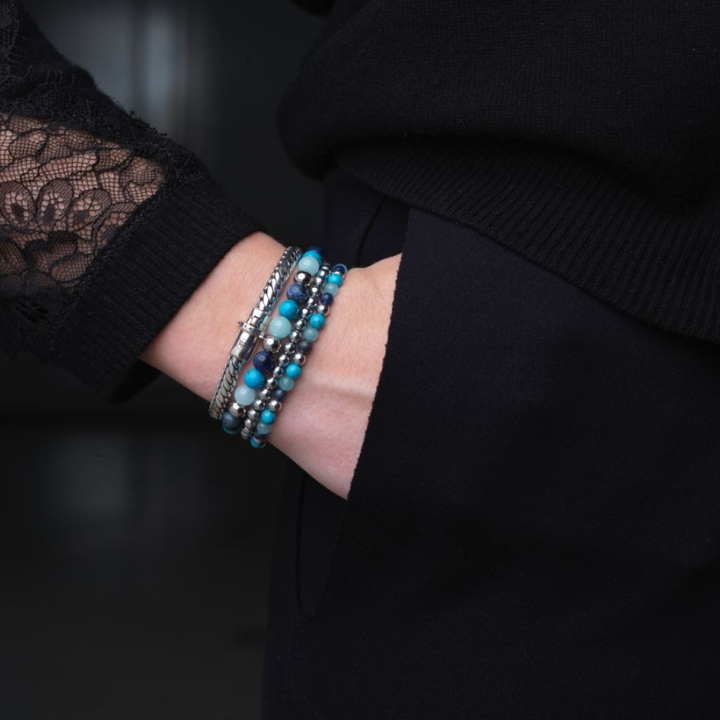 Een persoon in een top met zwarte kanten mouwen showt gestapelde blauwe, turquoise en zilveren kralenarmbanden met de stijlvolle Rebel & Rose Apollo Small six - 18,30 gr. 925 zilveren armband; de donkere achtergrond is onscherp.