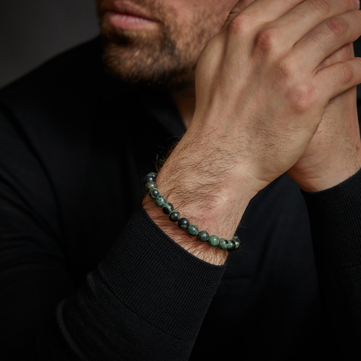 Een moderne man in een zwart shirt met lange mouwen laat zijn handen rusten en benadrukt de Rebel & Rose Green Rocks - 8mm Heren Kralen Armband om zijn pols. Zijn netjes getrimde baard en gedeeltelijk zichtbare gezicht benadrukken zijn stijlvolle look.