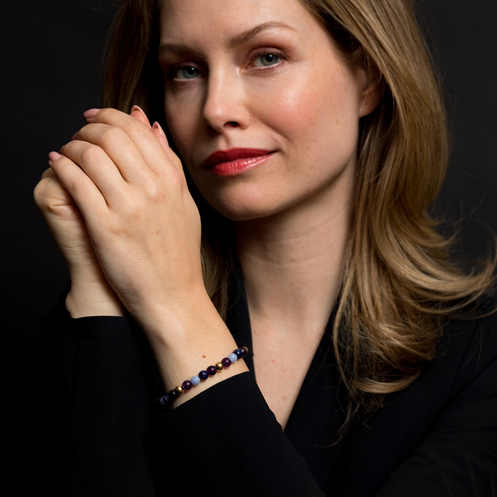 Een vrouw met lichtbruin haar en rode lippenstift poseert zelfverzekerd. Ze draagt een Rebel & Rose "Are Violets Blue?" armband Dames. 18 kt geelgouden geïoniseerde 6mm Kralen Armband Dames, gecombineerd met een zwart topje tegen een donkere achtergrond.