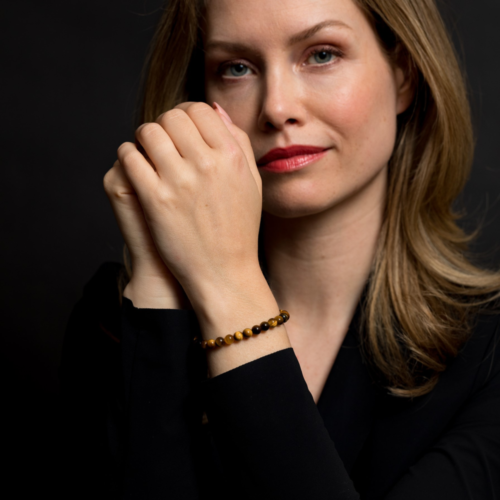 Een vrouw met lichtbruin haar en rode lippenstift poseert tegen een donkere achtergrond in het zwart met de Rebel & Rose Tiger Lily Gold - 6mm Kralen Armband Dames om haar pols, haar handen zachtjes gekruist in de buurt van haar gezicht.