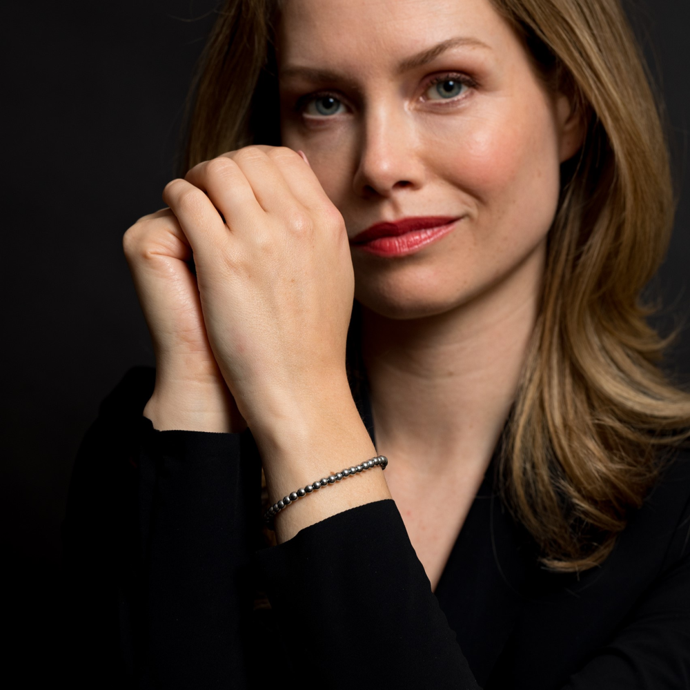 Een vrouw met lichtbruin haar en rode lippenstift poseert elegant tegen een donkere achtergrond, draagt een zwart topje en de Rebel & Rose Silver Shine - 4mm Kralen Armband Dames om haar pols, haar handen zachtjes geklemd bij haar gezicht.