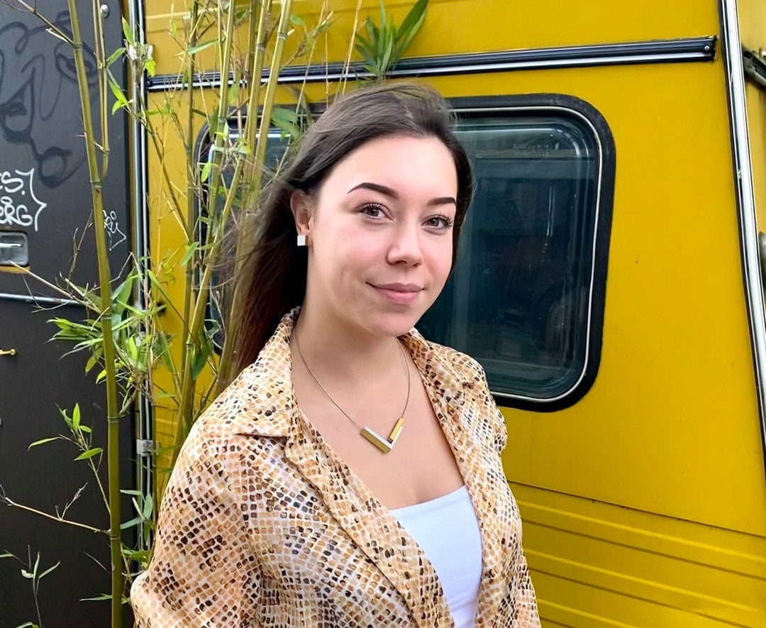 Een vrouw met lang bruin haar staat bij een geel voertuig, draagt een geprinte jas over een wit topje en een ketting van hypoallergeen chirurgisch staal. Groene planten staan op de achtergrond. (Let op: productgegevens zijn niet direct van toepassing op de scène.).