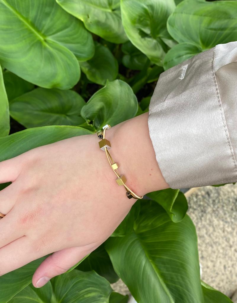 Een persoon die de CLIC Creations Armband speelse kubusjes goud A257G draagt, laat zijn hand rusten op weelderige groene bladeren. Ze zijn elegant gestyled in een lichtgekleurd satijnen shirt met geometrische gouden accenten.