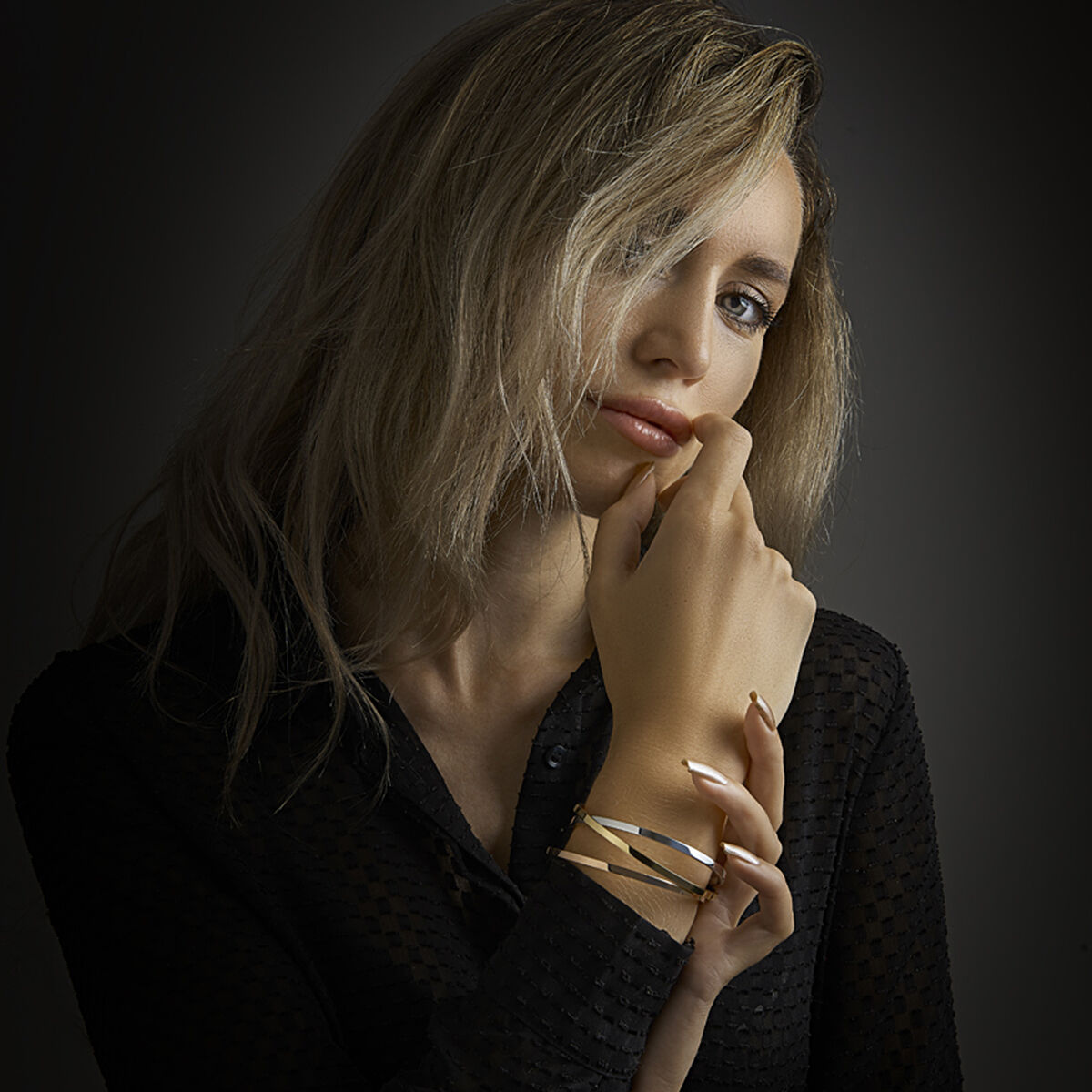 Een vrouw met golvend blond haar in een zwart topje met structuur kijkt naar de camera. Ze laat haar hand op haar kin rusten en draagt de armband scharnier massief vlakke buis 3 x 61 mm Zilgold van Van der Kooij Collectie om haar pols, tegen een donkere achtergrond.