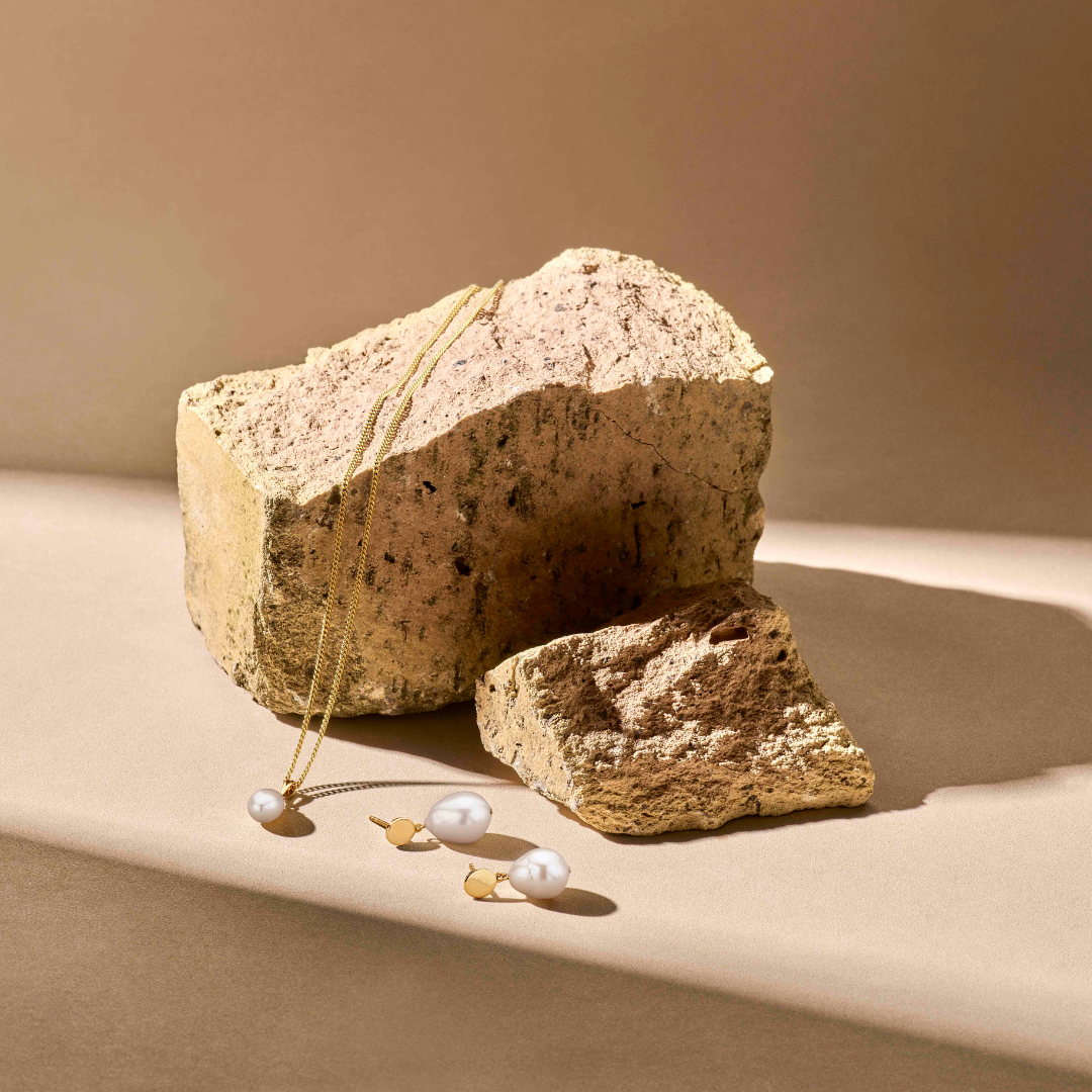 De Van der Kooij Collectie hanger parel 14K geelgoud en bijpassende oorbellen met grote zoetwaterparels liggen op een beige oppervlak naast lichtbruine stenen, terwijl zacht zonlicht hun elegante eikel benadrukt.