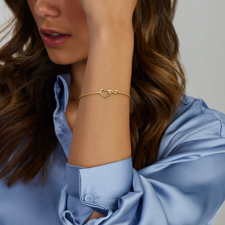Een vrouw met golvend bruin haar in een lichtblauw satijnen overhemd modelleert de Van der Kooij Collectie armband hart en infinity 16 + 3 cm 3 micron zilver verguld (geel), voorzien van een hart bedeltje- een elegante, relaxte look ideaal als Valentijn cadeau.