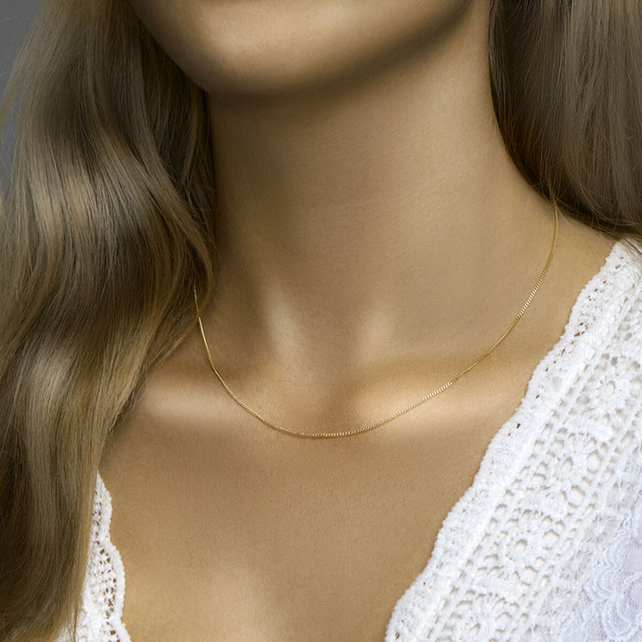 Een vrouw met lang lichtbruin haar draagt een Van der Kooij Collectie collier gourmette 4-zijdes geslepen 1,2 mm 41 + 4 cm 3 micron zilver verguld (geel) en een wit kanten topje, afgebeeld vanaf de schouders omhoog.