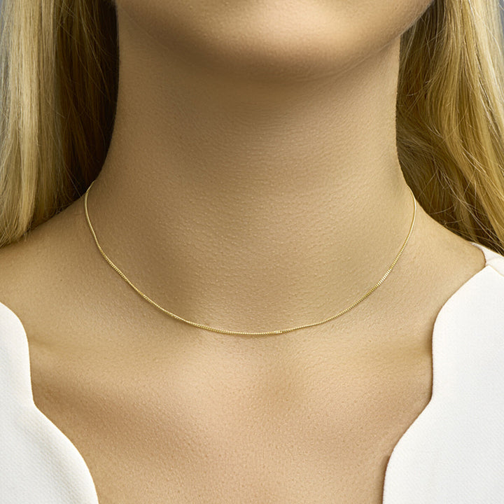 Een close-up van de hals van een vrouw met de Van der Kooij Collectie collier gourmette 1,0 mm 14K geelgoud choker. Ze heeft lang blond haar en is gekleed in een wit topje met een V-vormige halslijn.