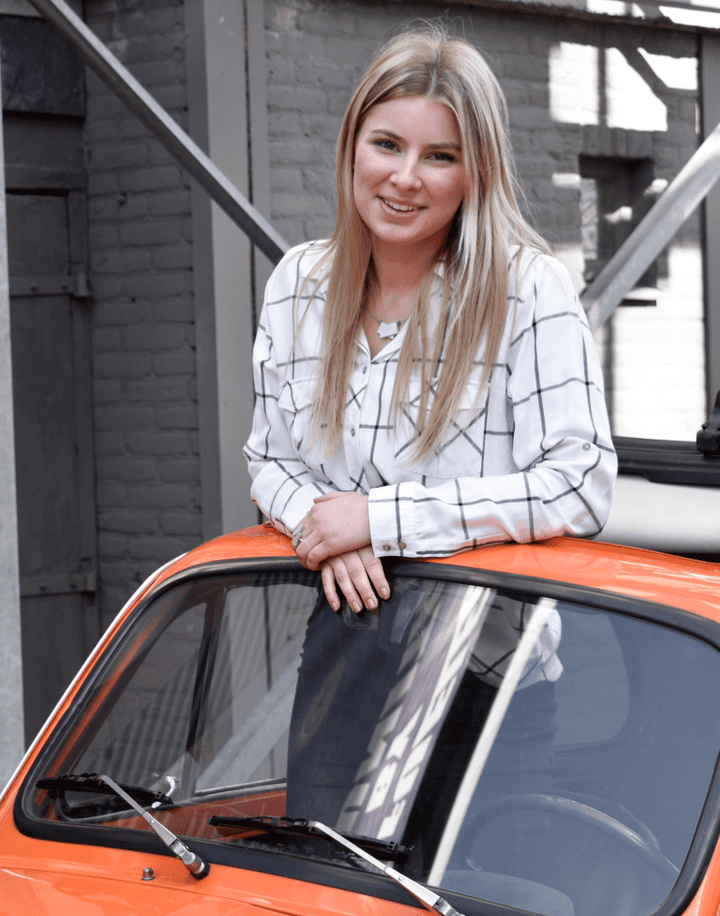 Een persoon met lang blond haar, gekleed in een wit geblokt shirt, lacht terwijl hij tegen de voorruit van een oranje auto leunt. Ze staan voor een bakstenen gebouw met metalen structuren, gekleed in CLIC Creations Collier gekruiste rechthoeken & hematiet C233.