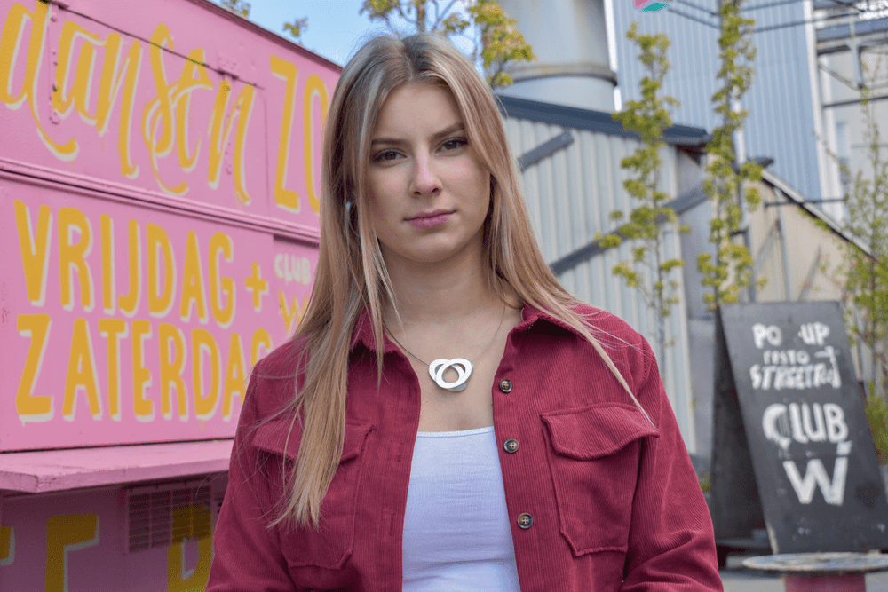 Een vrouw met lang blond haar draagt een rood shirt over een wit topje en staat voor een roze structuur met gele letters, wat het unieke ontwerp van CLIC Creations Collier Ovalen op elkaar C70, handgemaakt in Nederland, weerspiegelt. Borden en kleine bomen voegen stedelijke charme toe aan de achtergrond.