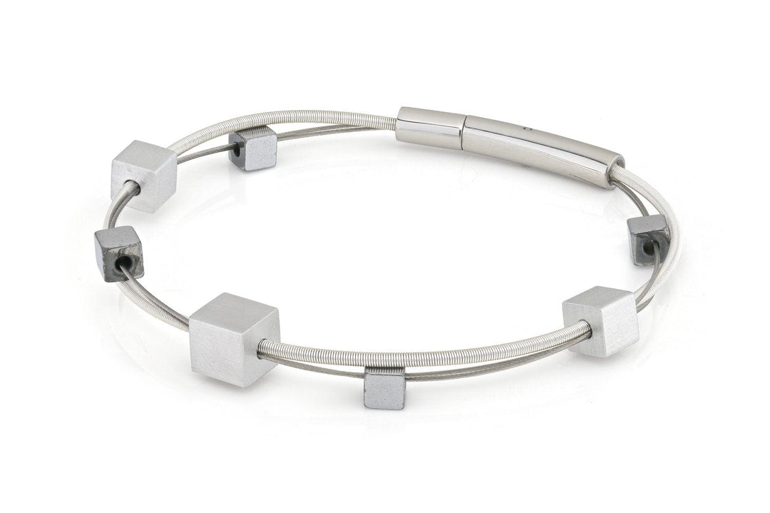De CLIC Creations Armband speelse kubusjes zilver A257 is een minimalistische zilveren armband, gemaakt van hypoallergene materialen, met een strak, dun draadontwerp versierd met gelijkmatig verdeelde vierkante metalen kralen en een cilindervormige sluiting.