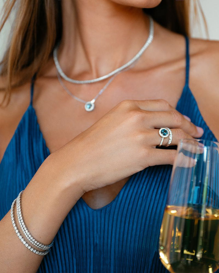 Een vrouw in een blauwe plooijurk houdt een glas witte wijn vast, draagt zilveren sieraden waaronder de Excellent Jewelry 14 karaat witgouden tenniscollier met 146 diamanten (2,81 crt), plus een versierde ring en gestapelde armbanden.