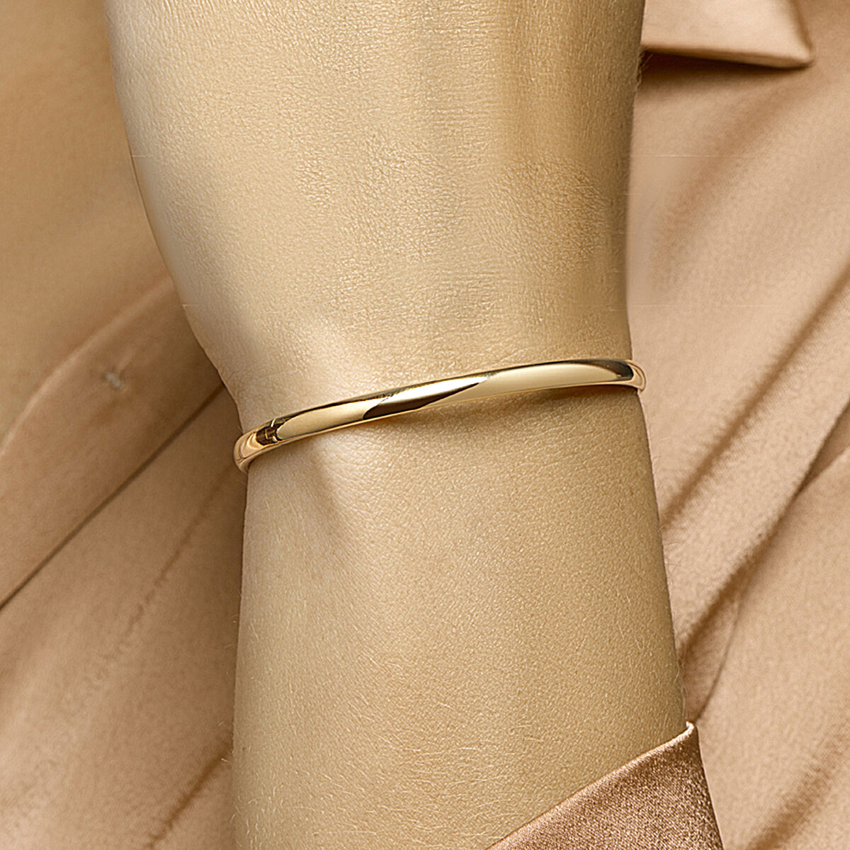 Een close-up van een Van der Kooij Collectie bangle scharnier massief ovale buis 4,0 mm Zilgold (geelgoud met zilverkern) gedragen om de pols over een beige satijnen shirt, met bijpassende achtergrond die het glanzende, eenvoudige ontwerp laat zien.