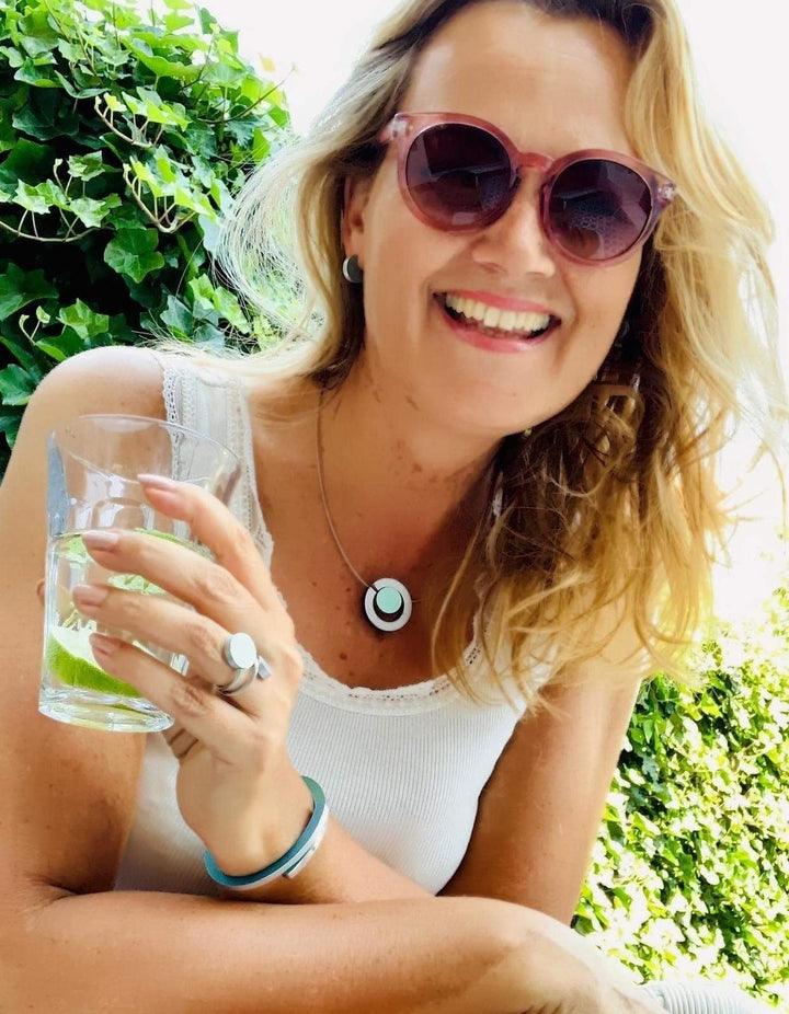 Een lachende vrouw met lang blond haar en een zonnebril draagt een uniek ontworpen wit tanktopje en CLIC Creations Ring Twee ronde vormen Roze R5RO, terwijl ze een glas limoenwater vasthoudt. Handgemaakt in Nederland, ze staat buiten met weelderig groen gebladerte achter haar.