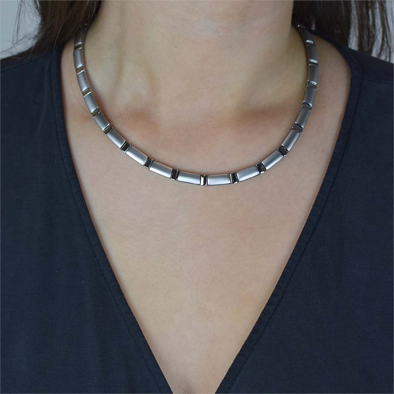 Een close-up toont iemand die de Boccia Titanium Collier 0845-01 draagt, een zilveren, matte ketting met een gesegmenteerd ontwerp. Het individu is gekleed in een donkere top met V-hals tegen een onopvallende achtergrond.