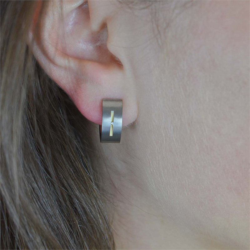 Close-up van een persoon met steil bruin haar en een lichte huid die Boccia Titanium Oorringen Bicolor dia2/0.01 - 0521-03 draagt: elegante zilveren oorringen met een klein verticaal gouden accent van Boccia.