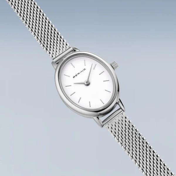 De Bering 11020-004 is een zilverkleurig dameshorloge met een witte ovalen wijzerplaat, een minimalistisch ontwerp, een metalen band van mesh en eenvoudige uurmarkeringen tegen een lichtblauwe achtergrond met kleurverloop.
