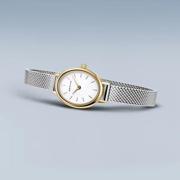 De Bering 11020-010 is een zilvergaas dameshorloge met een witte ovale wijzerplaat, gouden lunette, minimalistische uurmarkeringen, duurzaam saffierglas en nauwkeurig quartz uurwerk tegen een lichtblauwe gradiënt achtergrond.
