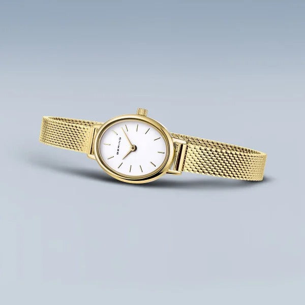 Het Bering dameshorloge 11020-334 heeft een goudkleurige mesh band, witte ovale wijzerplaat met minimalistische zwarte uurmarkeringen en twee wijzers, kwarts uurwerk, gepresenteerd op een lichtblauwe achtergrond.
