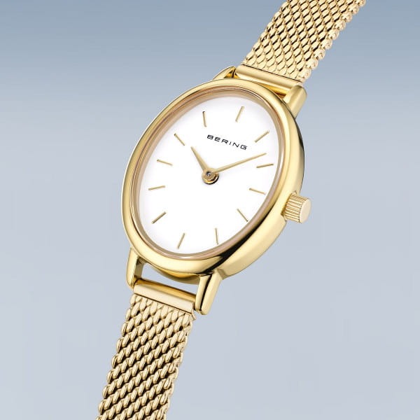 Het Bering dameshorloge 11020-334 heeft een goudkleurige mesh band, quartz uurwerk en witte wijzerplaat met gouden tijdsaanduiding, en is afgebeeld tegen een lichtblauwe achtergrond.