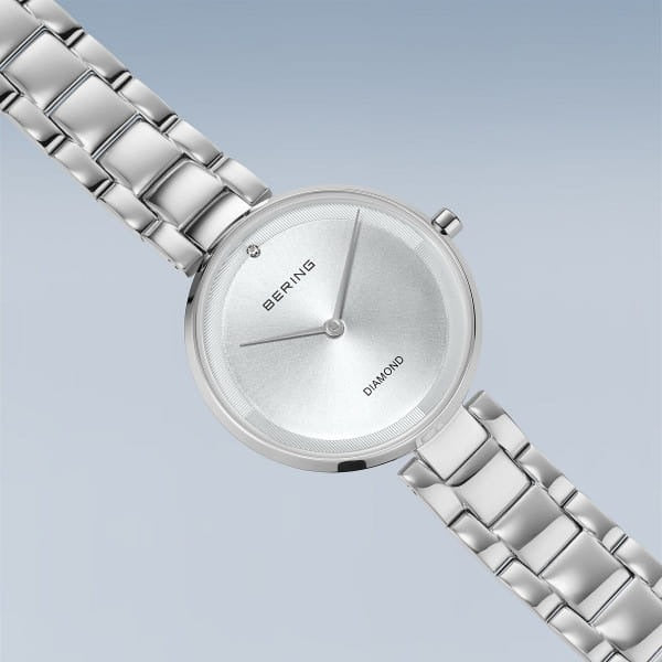 De Bering 17529-700 dameshorloge heeft een minimalistische ronde wijzerplaat met saffierglas, een enkele diamant op 12 uur en een zilveren schakelarmband, weergegeven op een lichtblauwe gradiënt achtergrond.