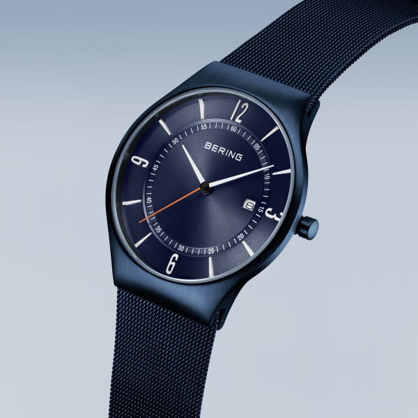 De Bering 15940-393 is een stijlvol herenhorloge uit de Classic collectie met een donkerblauwe mesh band, minimalistische wijzerplaat, witte uurmarkeringen, datumvenster en dunne rode secondewijzer op een lichtblauwe gradiënt achtergrond.
