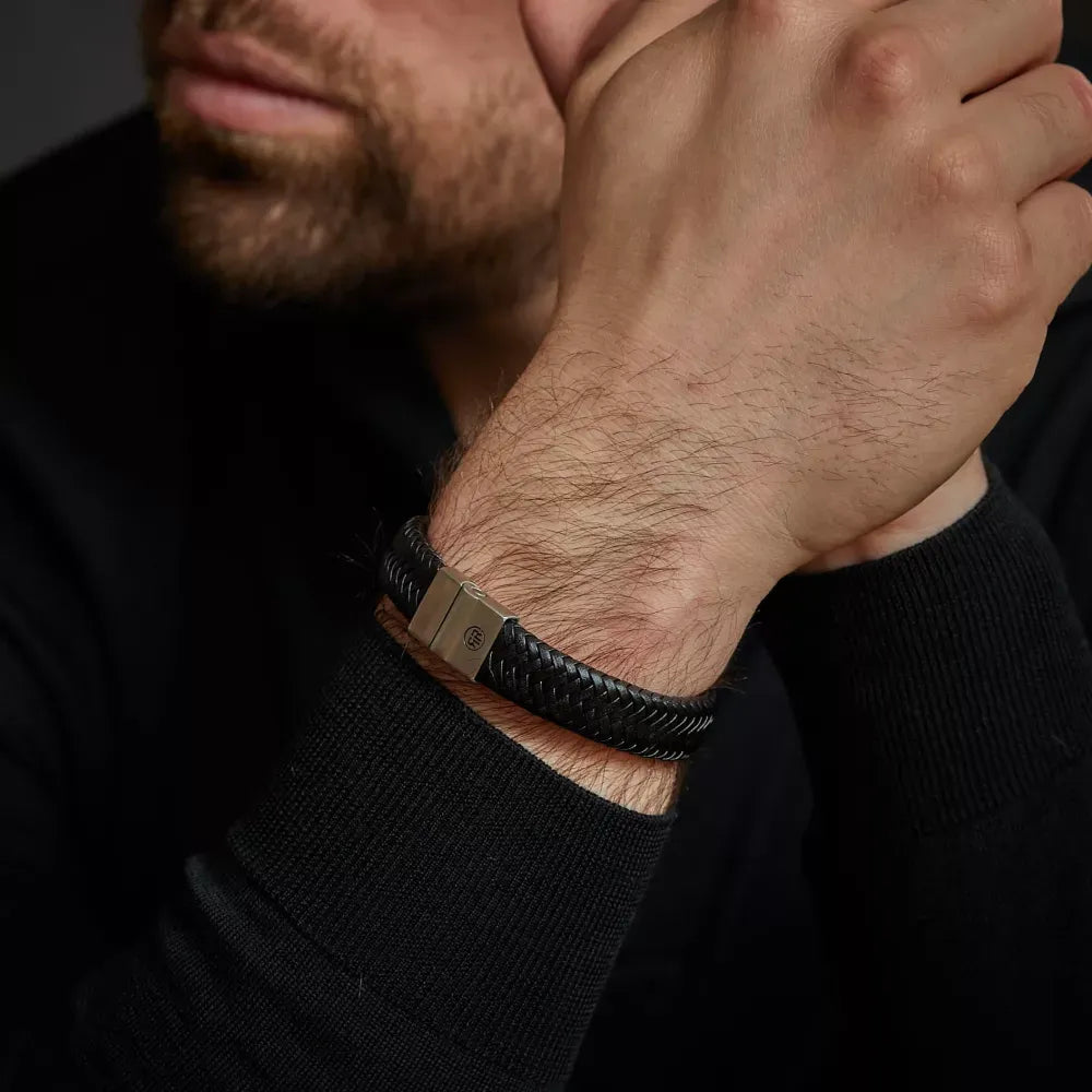 Een bebaarde man in een zwart shirt met lange mouwen draagt de Full Metal Plaid Black Matt 15 mm gevlochten leren armband van Rebel & Rose met een metalen sluiting. Hij laat zijn kin op zijn hand rusten tegen een donkere achtergrond.