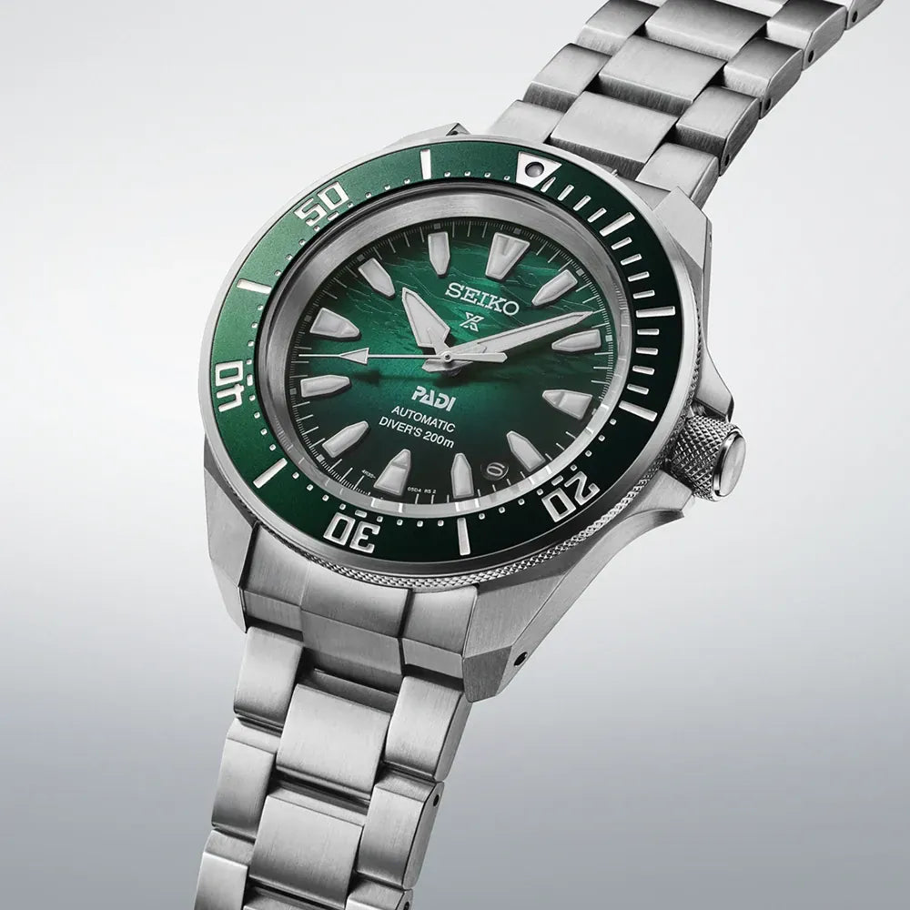Het Seiko heren horloge SRPL53K1 heeft een groene bezel en wijzerplaat, zilveren indexen en wijzers, en een stalen band. Dit stijlvolle Seiko horloge wordt diagonaal getoond op een lichte achtergrond.