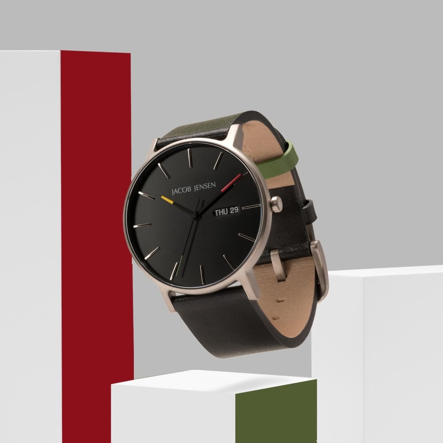 De Jacob Jensen 162 is een strak, modern polshorloge met een zwarte wijzerplaat en een zilveren titanium kast. Het heeft een quartz uurwerk, een zwarte leren band en slanke zilveren uurmarkeringen, allemaal afgebeeld op een geometrische minimalistische achtergrond.