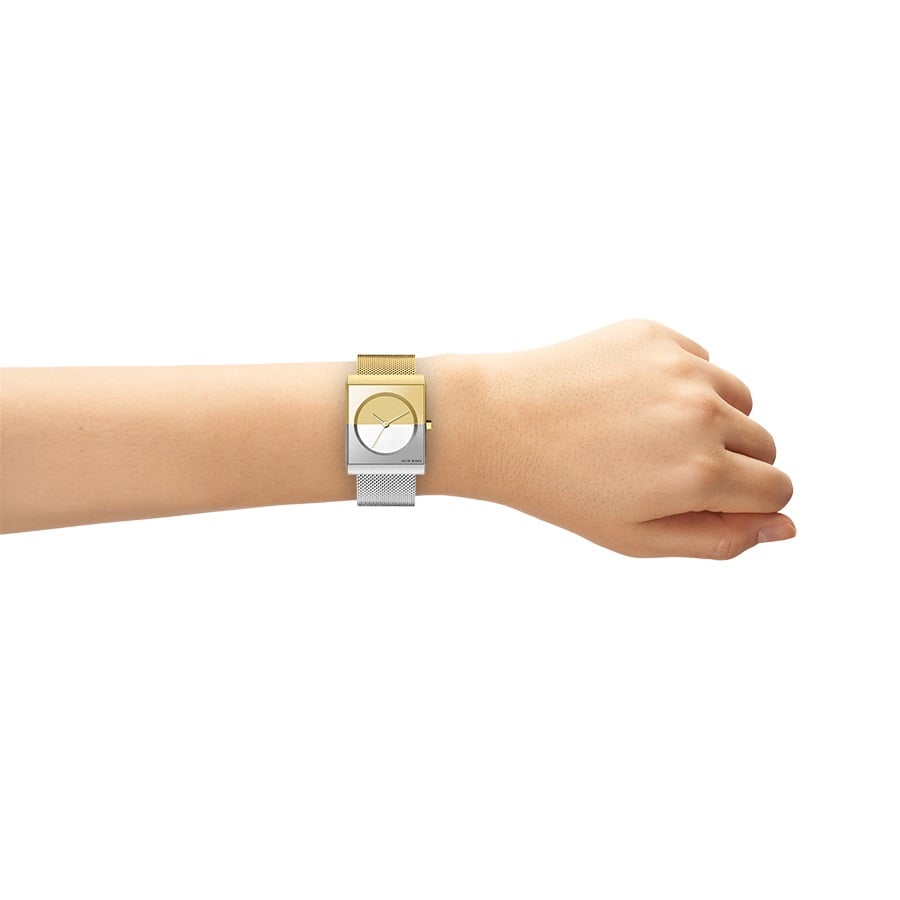 Een hand draagt een Jacob Jensen 526 horloge met een tweekleurige metalen band en een rechthoekige wijzerplaat. Het bovenste deel van de quartz wijzerplaat is geel en het onderste deel is zilver met saffieraccenten, tegen een effen witte achtergrond.