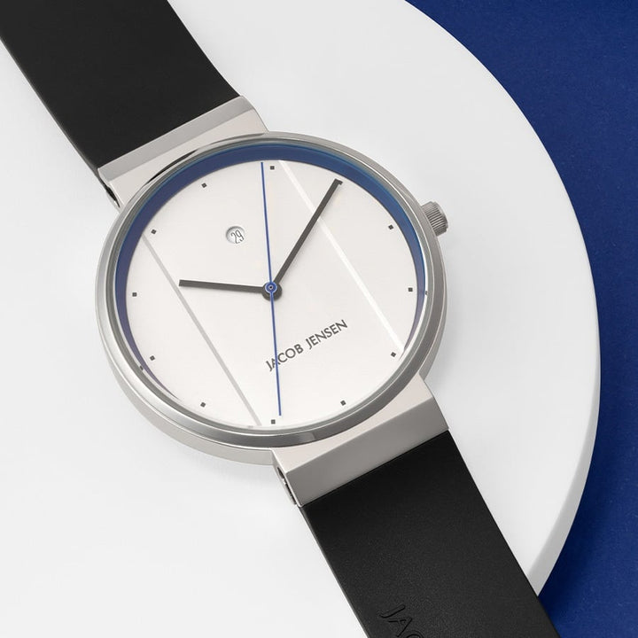 Het Model 750 polshorloge van Jacob Jensen combineert minimalistisch design met een quartz uurwerk, met een roestvrijstalen kast, zwarte band en een witte wijzerplaat met dunne zwarte uurmarkeringen en een blauwe secondewijzer. Het wordt weergegeven op een rond, wit oppervlak tegen een blauwe achtergrond.