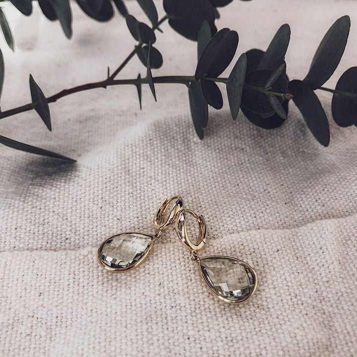 De Stralende Geelgouden Oorringen met Citrien Aanhangers van Excellent Jewelry liggen op textuur crème stof, met een eucalyptus tak diagonaal op de achtergrond.